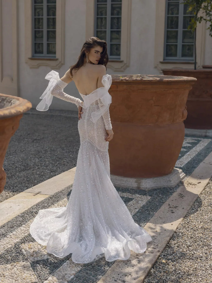 A woman wearing a white lace mermaid-style wedding dress with sheer flutter sleeves and an open back, standing outdoors in front of a building with large terracotta planters.