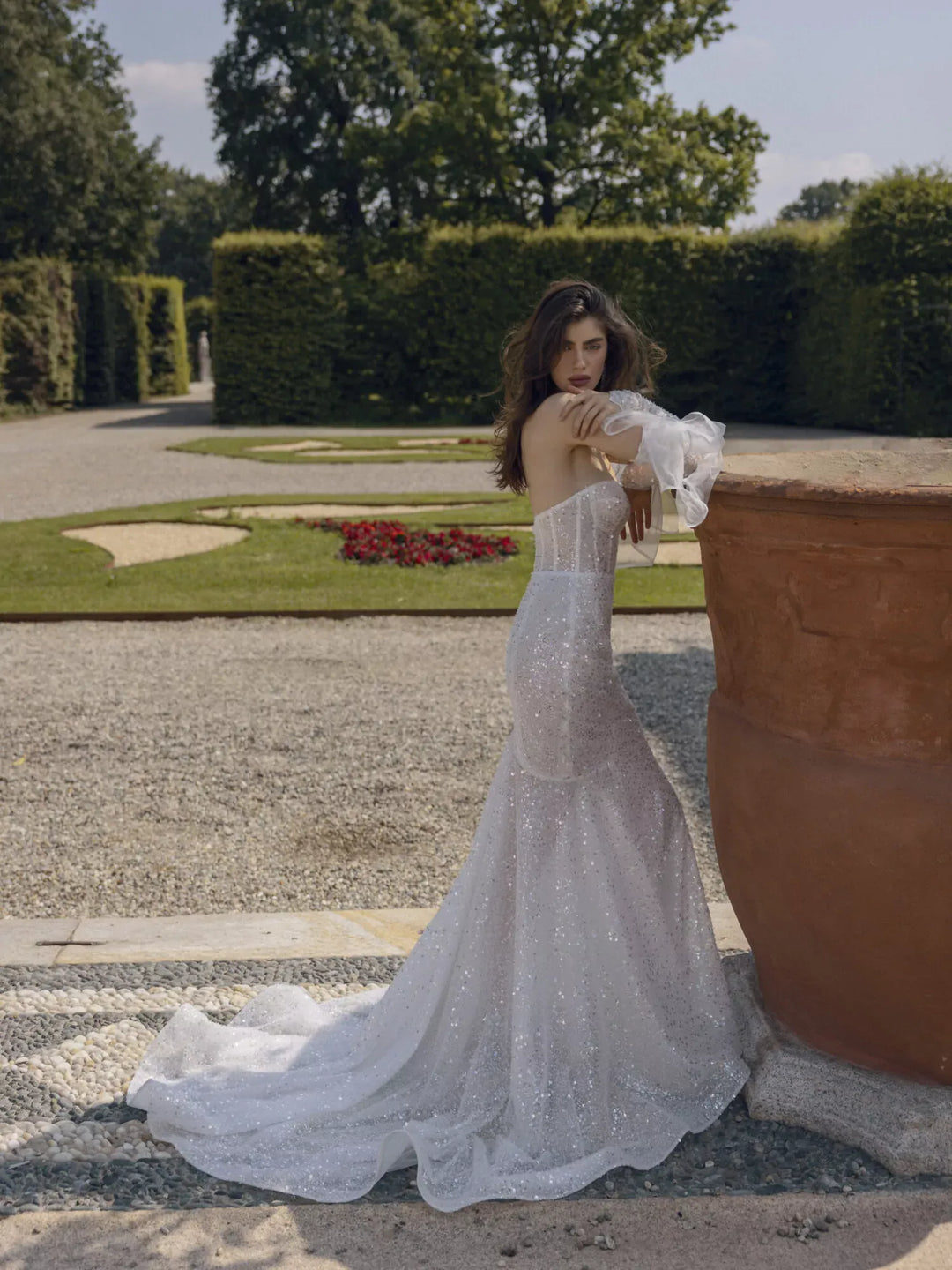 A woman wearing a sparkling white strapless mermaid gown with ruffled sleeves stands beside a large terracotta planter in a formal garden setting.