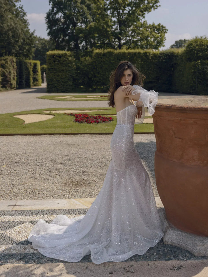 A woman wearing a sparkling white strapless mermaid gown with ruffled sleeves stands beside a large terracotta planter in a formal garden setting.