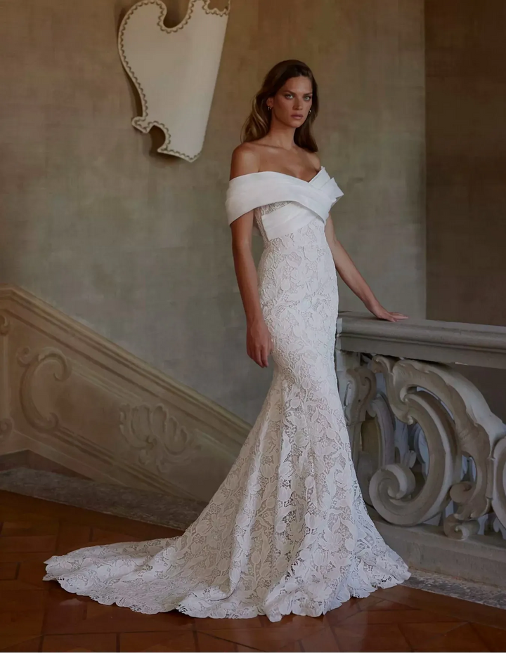 Woman in a white off-shoulder lace wedding dress standing in an elegant interior setting.
