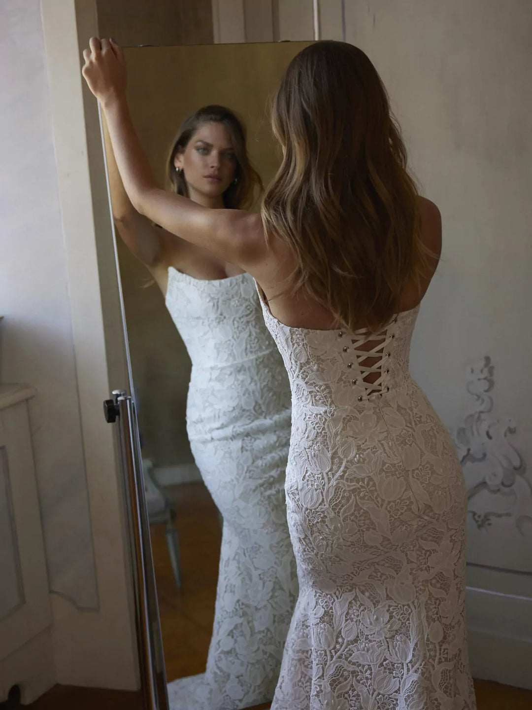Woman in a white lace wedding dress looking at herself in a mirror.