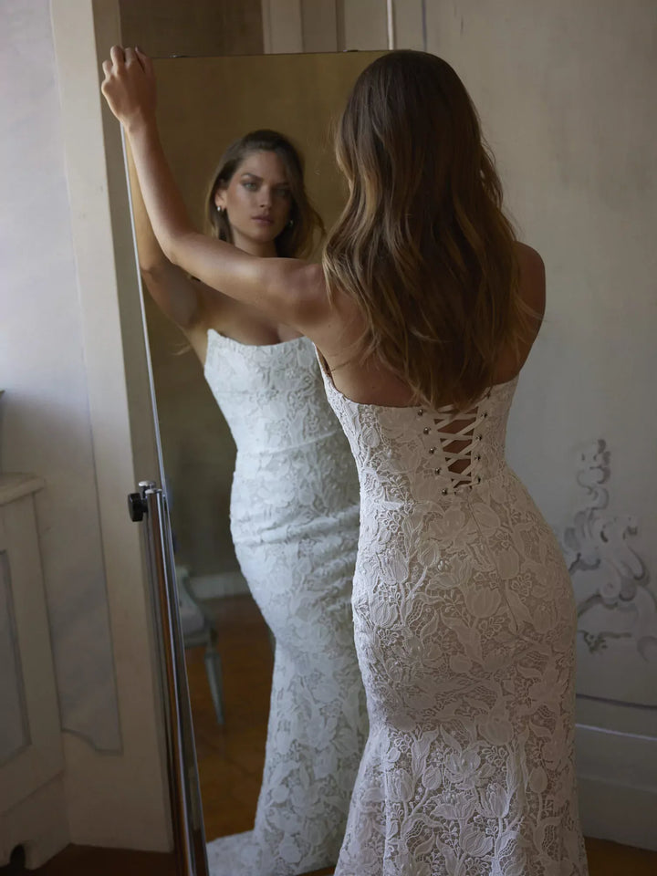 Woman in a white lace wedding dress looking at herself in a mirror.