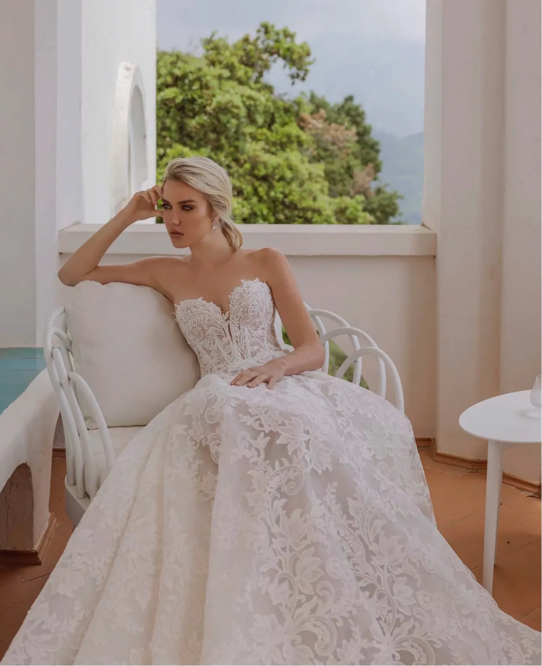 Woman in a white lace wedding dress sitting on a balcony with a scenic view.