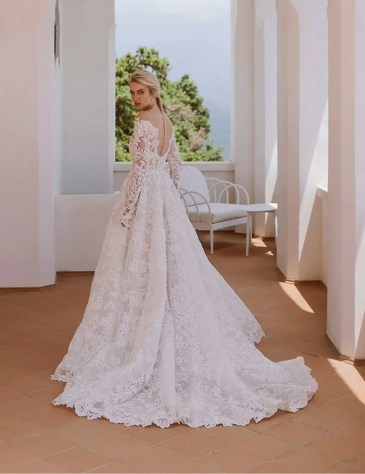 Woman in a white lace wedding dress standing in a sunlit room with arched windows.