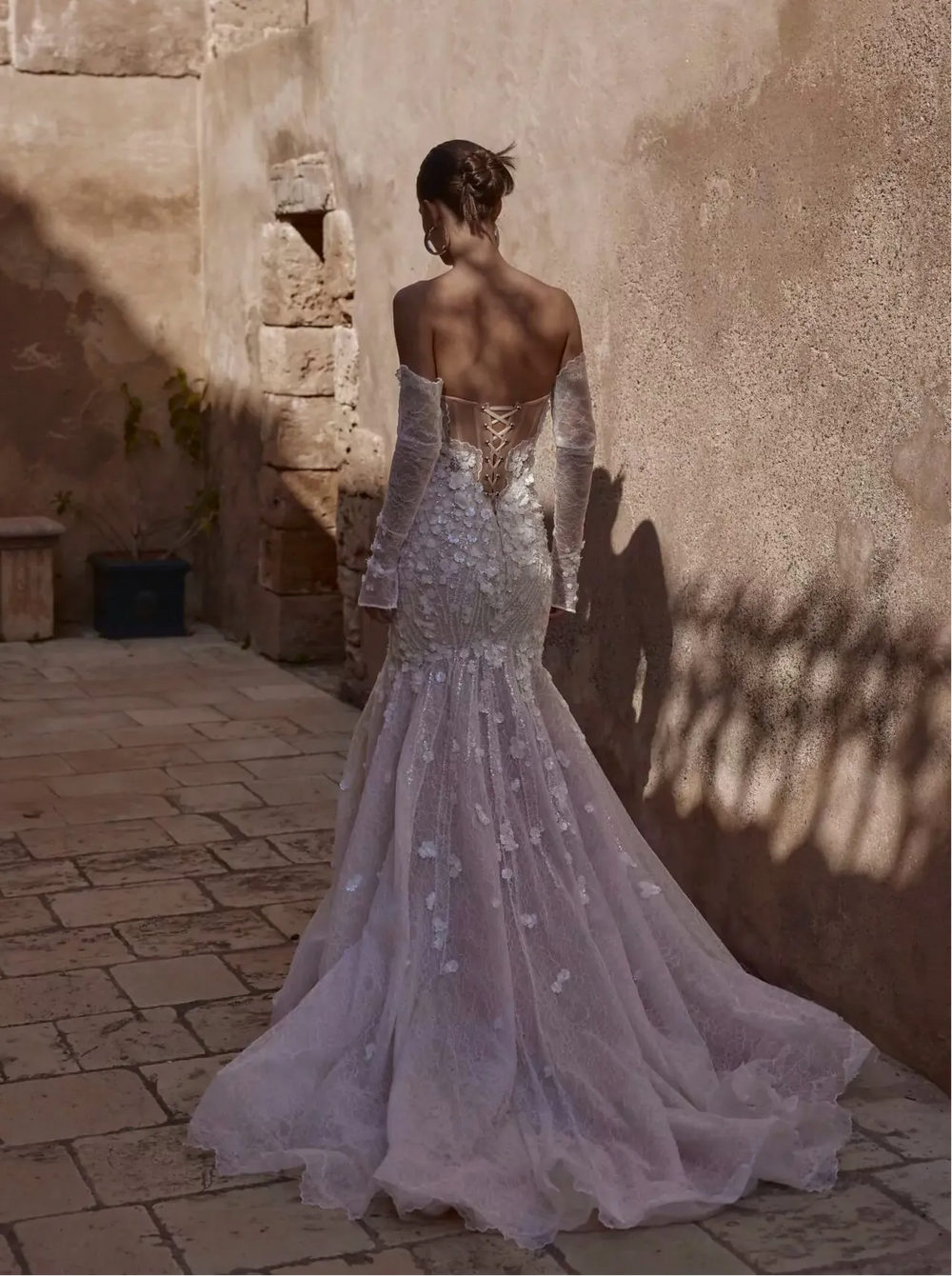 Woman in a sparkling silver dress standing in a sunlit outdoor setting with stone walls.