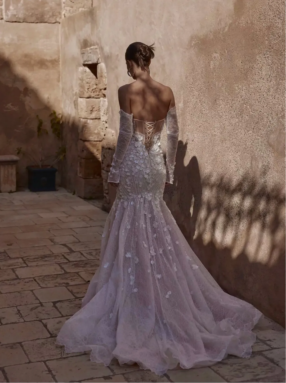 Woman in a sparkling silver dress standing in a sunlit outdoor setting with stone walls.