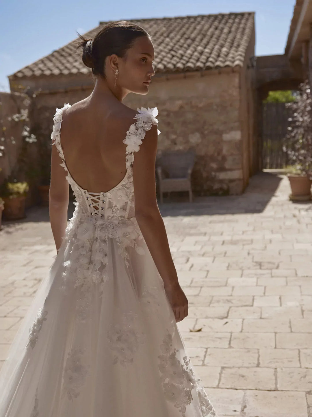 Woman in a white lace wedding dress standing outdoors with stone building and potted plants in the background.