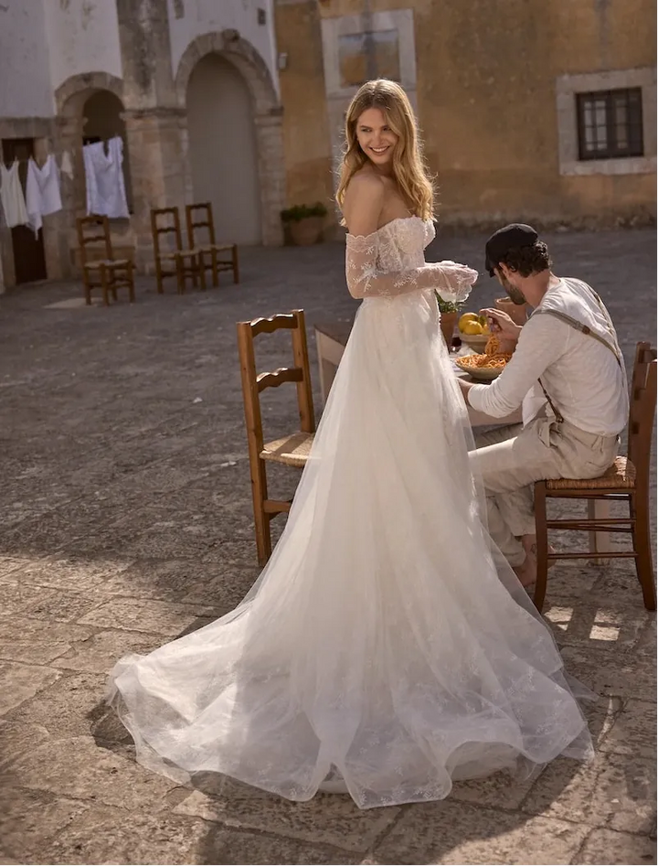 A bride in a long off-the-shoulder tulle wedding gown stands in a sunlit stone courtyard, while a man sits at a table nearby. The setting includes rustic buildings, chairs, and warm natural light.