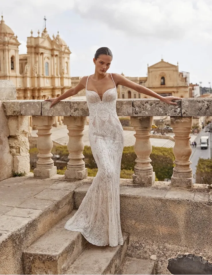 A woman wearing a fitted white lace wedding gown stands on stone steps outdoors, leaning on a balustrade with historic buildings and a cloudy sky in the background.