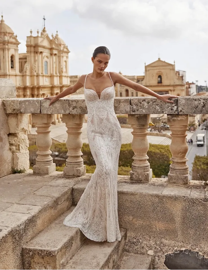 A woman wearing a fitted white lace wedding gown stands on stone steps outdoors, leaning on a balustrade with historic buildings and a cloudy sky in the background.