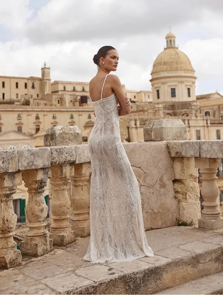 A woman wearing a sleeveless lace wedding gown stands on a stone terrace with a historic domed building in the background under a cloudy sky.
