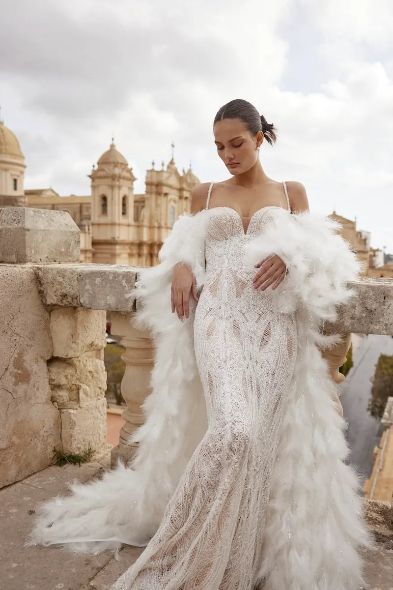 A woman wearing a white lace mermaid-style wedding gown with a voluminous feathered overskirt stands outdoors on a stone terrace with a historic building in the background under a cloudy sky.