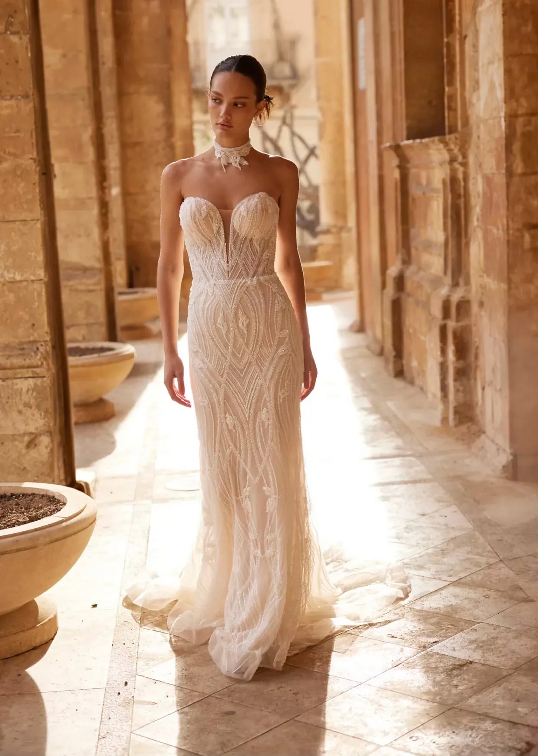 Woman in a white lace wedding dress standing in a sunlit stone corridor.