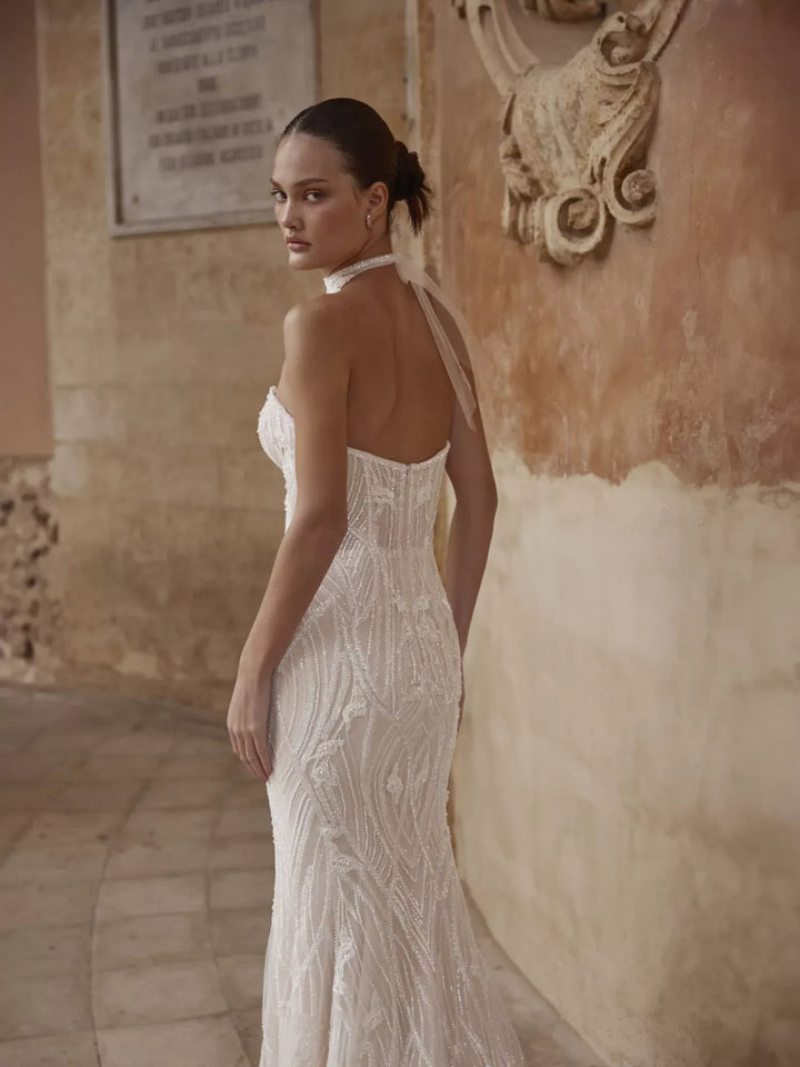 Woman in a white evening gown with a textured back design, standing against an ornate wall.