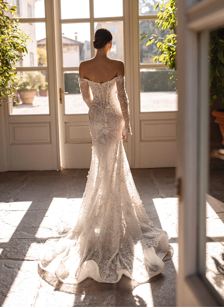Woman in a sparkling wedding dress standing in a sunlit room with large windows.