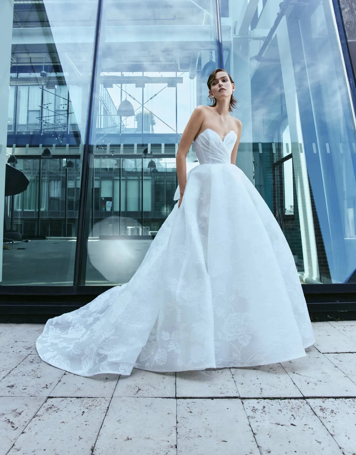 A woman wearing a white strapless wedding gown with a sweetheart neckline and a full ball gown skirt stands outdoors in front of a modern glass building.