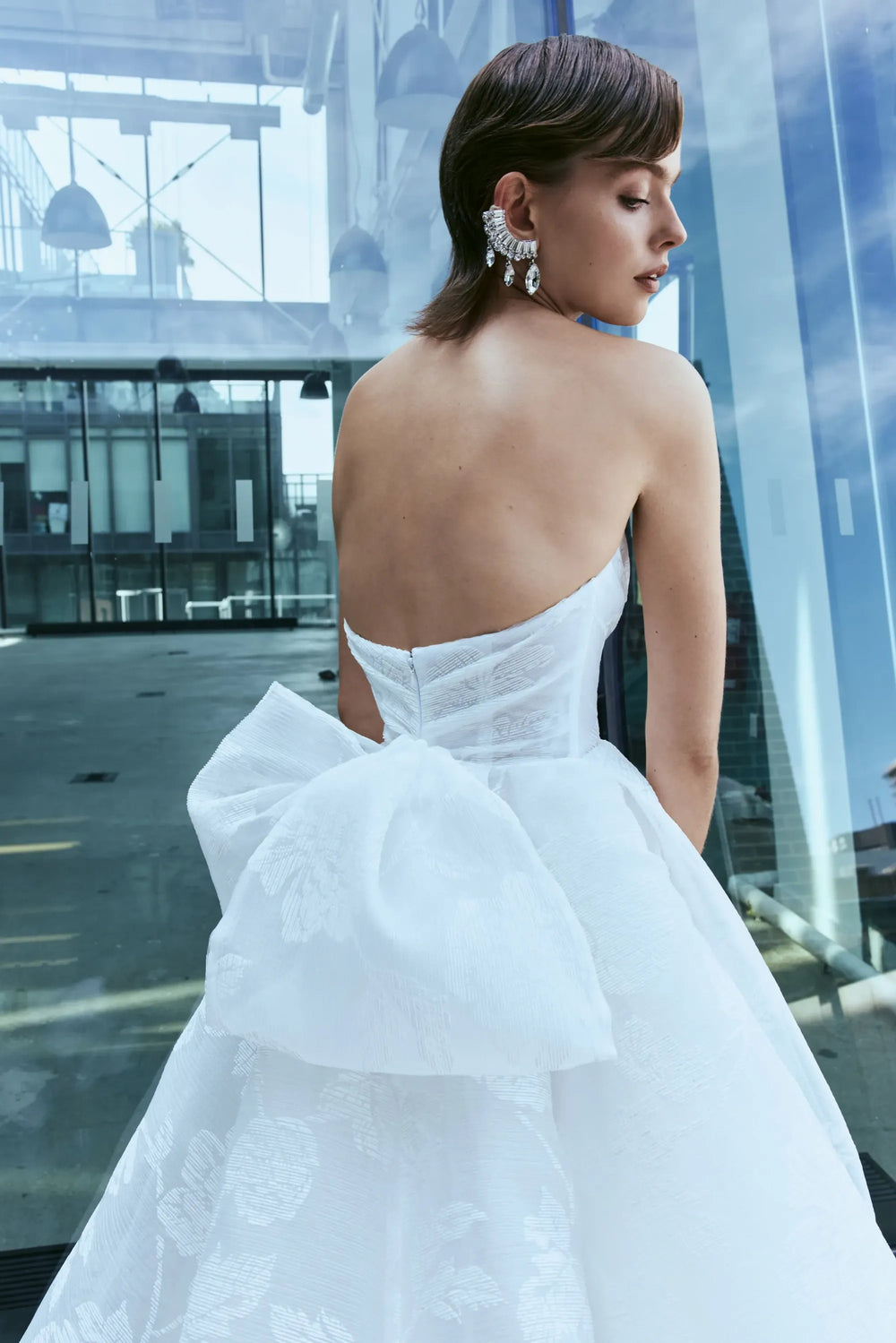 A woman wearing a strapless white ballgown with a large bow at the back, photographed from behind in a modern glass-walled interior.