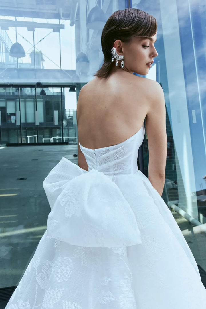 A woman wearing a strapless white ballgown with a large bow at the back, photographed from behind in a modern glass-walled interior.