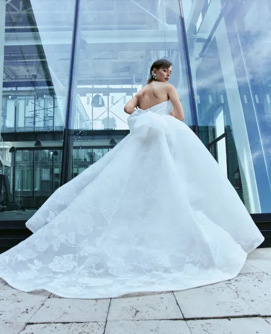 A woman wearing a voluminous white wedding gown stands in front of a modern glass building, holding the train out wide. The dress has a strapless one-shoulder design with a fitted bodice and a very full skirt with floral lace embroidery.