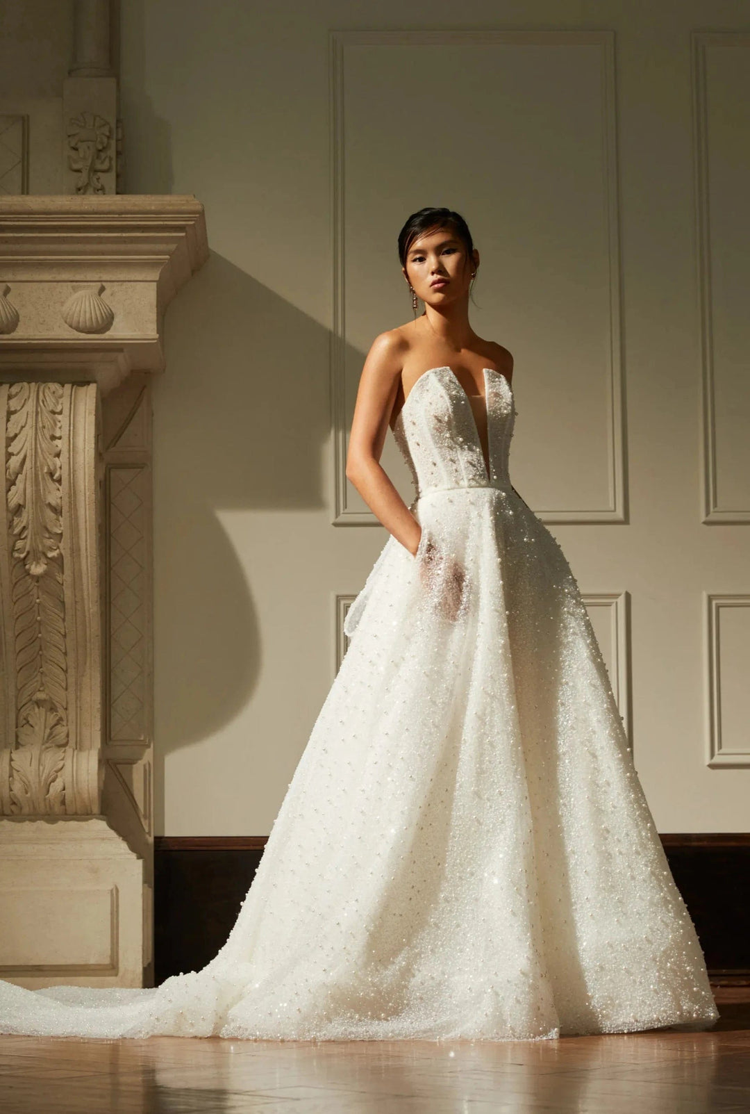 A woman modeling a strapless white wedding gown with a fitted bodice, sheer lace overlay, and a full textured skirt with a long train, standing in a sunlit interior with paneled walls and a carved fireplace.
