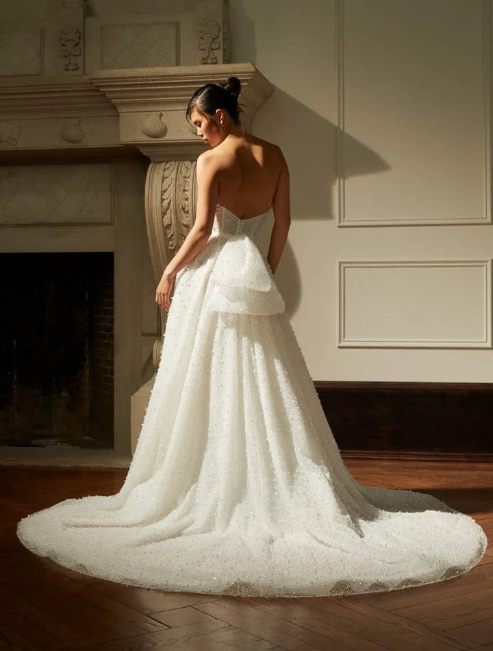 A woman wearing a white wedding gown stands with her back to the camera in an elegant room with a fireplace and wood floor. The dress has a structured lace bodice, a layered ruffled overlay, and a long flowing train.