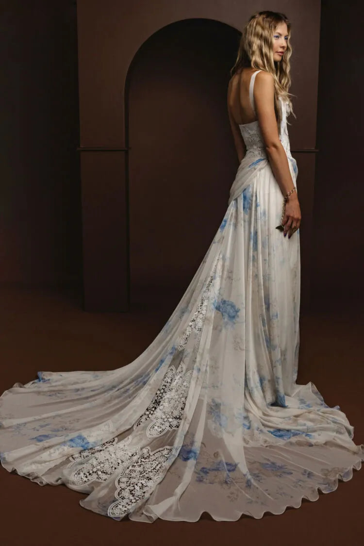 A woman modeling a floor-length white gown with blue floral print and lace trim, photographed in a dark brown studio setting with an arched backdrop.