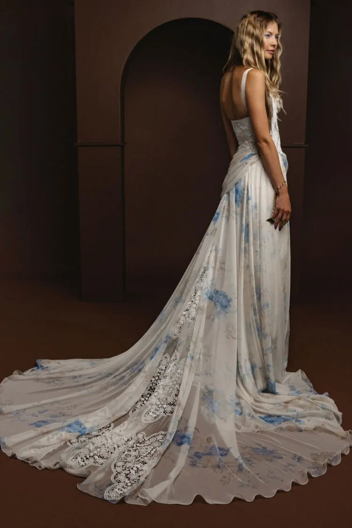 A woman modeling a floor-length white gown with blue floral print and lace trim, photographed in a dark brown studio setting with an arched backdrop.