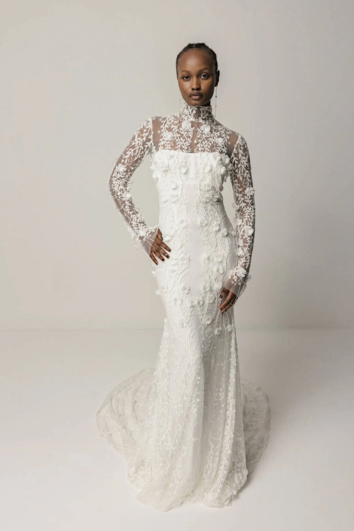 Model wearing a white wedding dress with floral details on a plain background