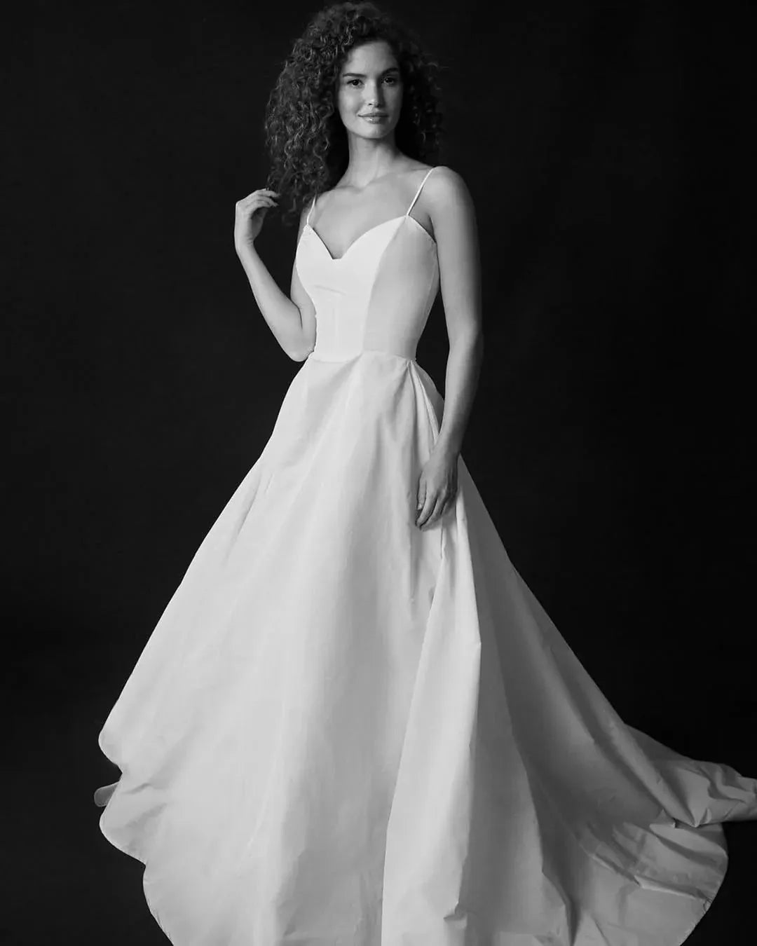 A woman modeling a sleeveless white A-line wedding gown with thin spaghetti straps, a sweetheart neckline, and a full flowing skirt against a dark studio background.
