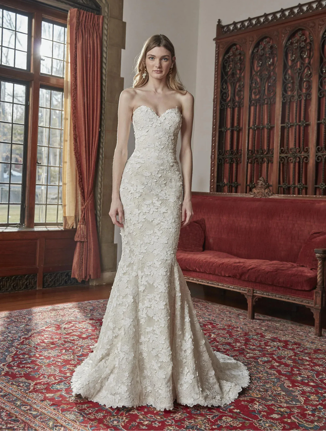 Woman in a strapless lace wedding dress standing in an elegant room with red curtains and a red sofa.