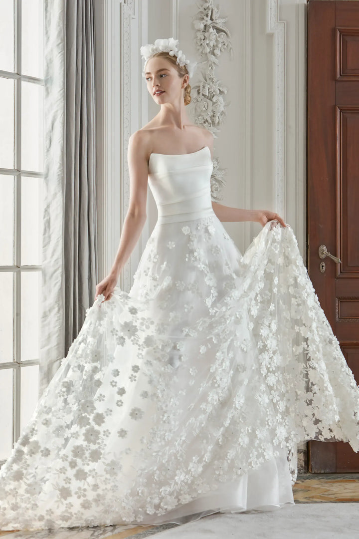 Woman wearing a white strapless wedding dress with floral details in a room with large windows.
