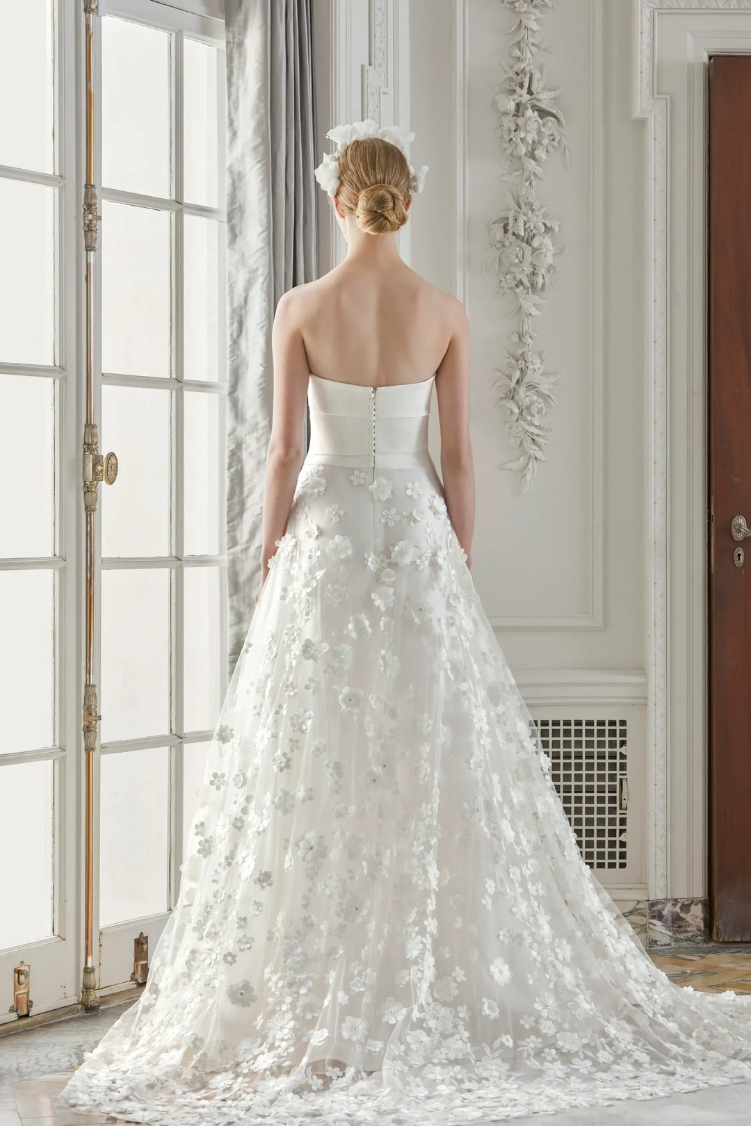Woman in a white wedding dress standing in a room with large windows.