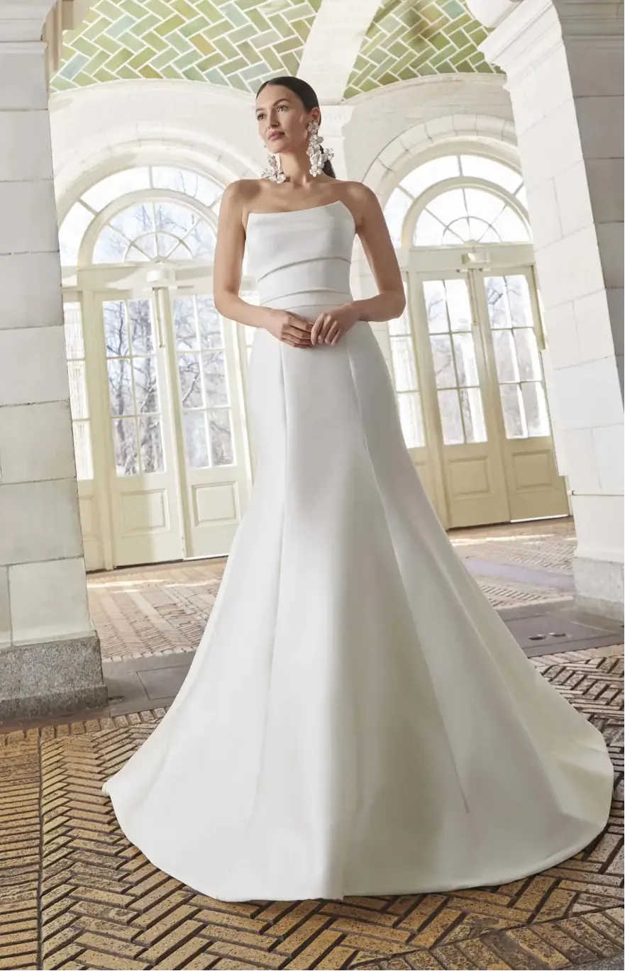 Woman in a white strapless gown standing in an elegant interior setting with large windows.