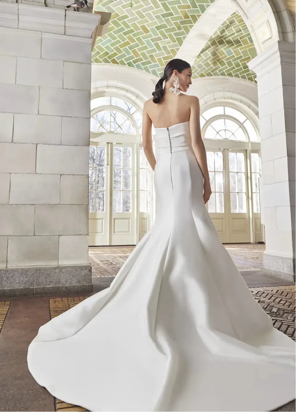 Woman in a white wedding dress standing in an elegant indoor setting with arched windows.