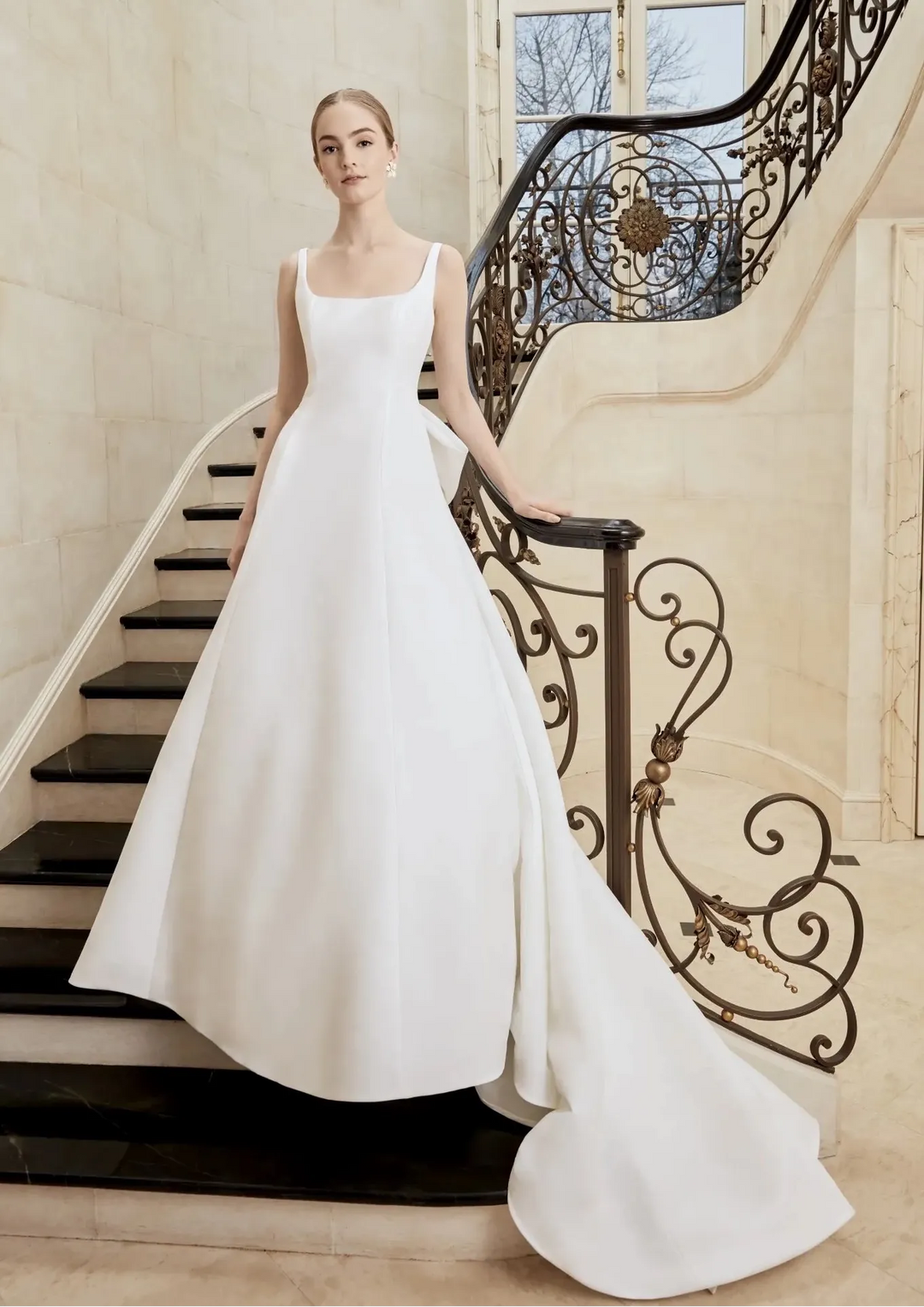 A woman wearing a sleeveless white A-line wedding gown stands on an indoor staircase with a dark wrought-iron railing, holding the handrail. The dress has a straight neckline, thin straps, and a full skirt with a long train.