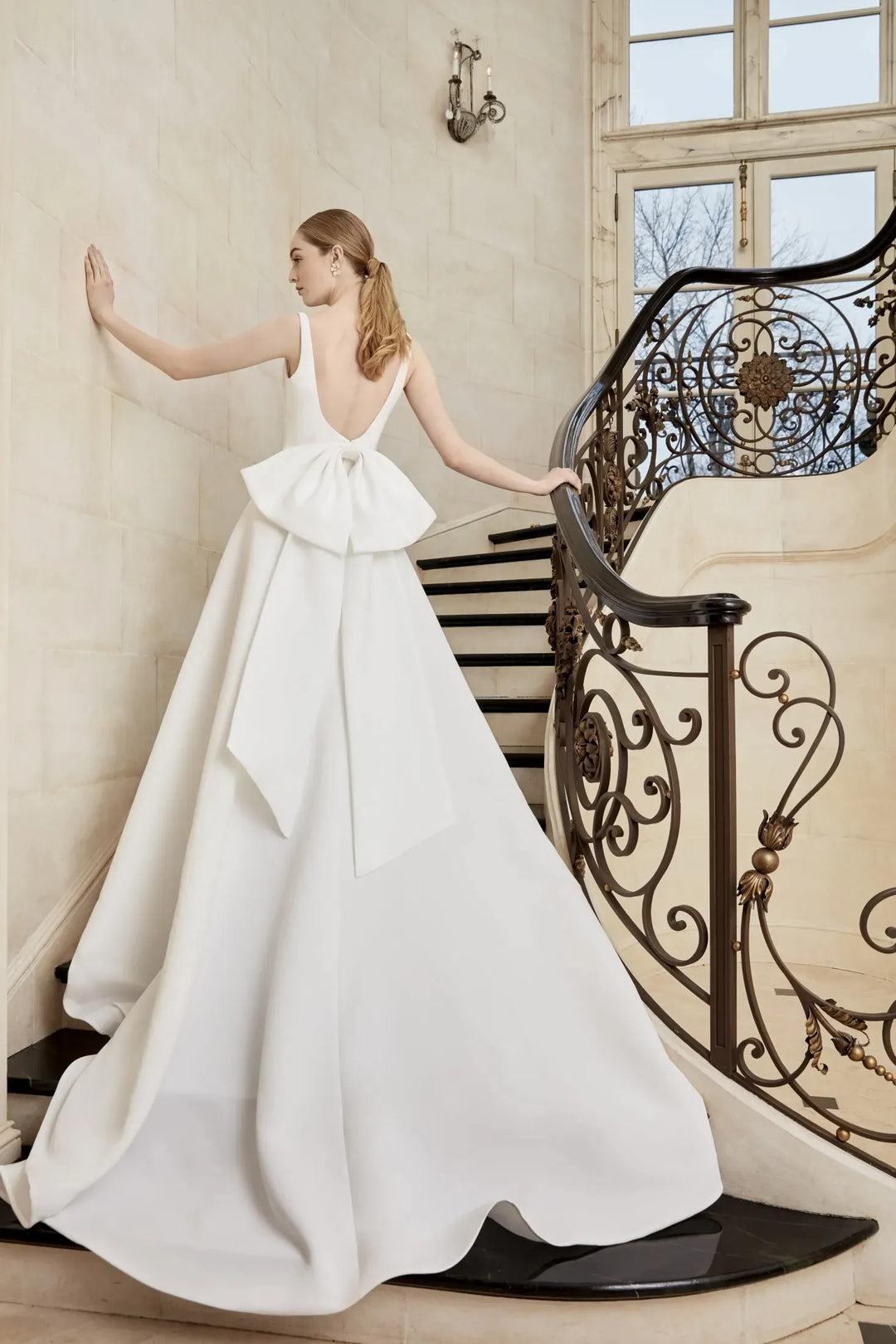 A woman wearing a white wedding gown with a large bow at the waist, standing on an indoor staircase with a decorative iron railing.