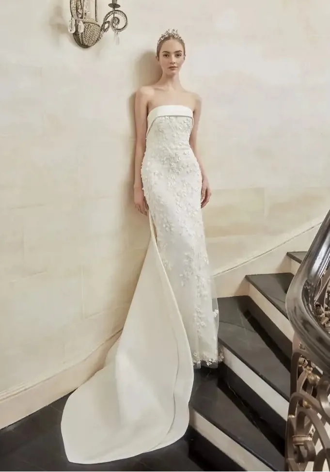 A woman wearing a strapless white wedding gown with floral lace applique and a long draped train, standing on an indoor staircase against a cream wall.