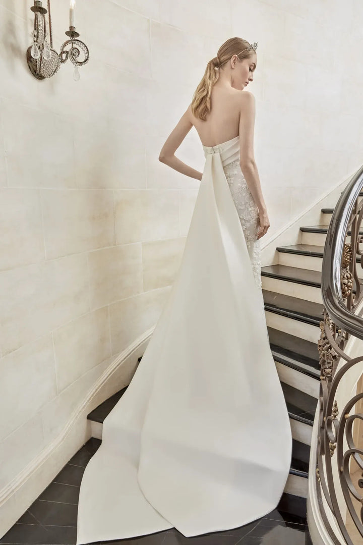 A woman wearing a strapless ivory wedding gown with a dramatic train, standing on a staircase in a grand interior.