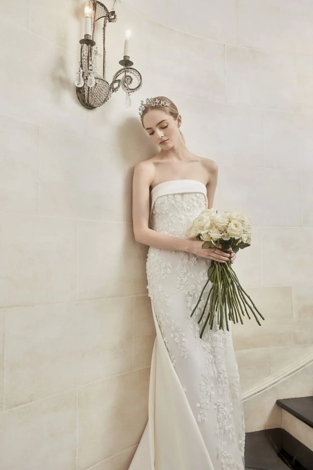 A woman in a strapless white lace wedding gown stands against a cream stone wall, holding a bouquet of white flowers. A lit wall sconce is visible above her.