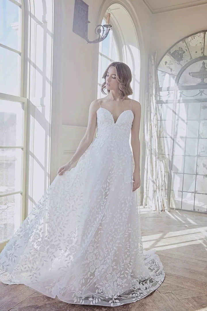 A woman wearing a strapless white lace wedding gown stands in a sunlit room with tall arched windows and a wood floor.