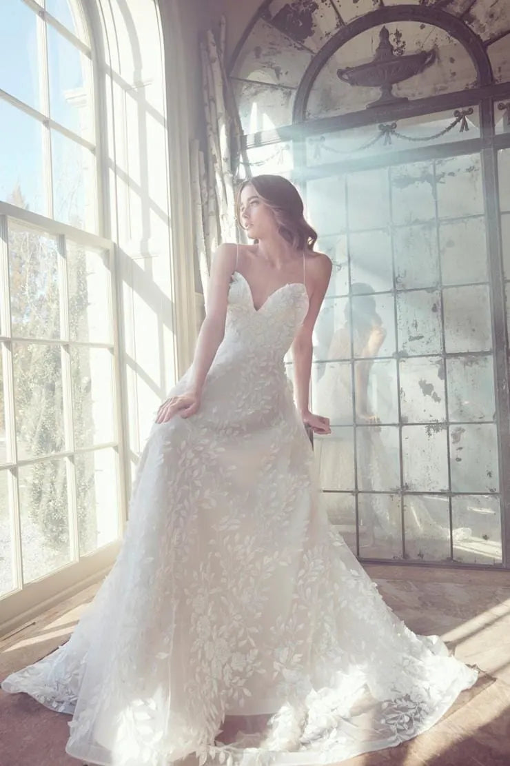 A woman wearing a strapless white lace wedding gown stands in a sunlit room with large arched windows and a mirror reflecting her silhouette.