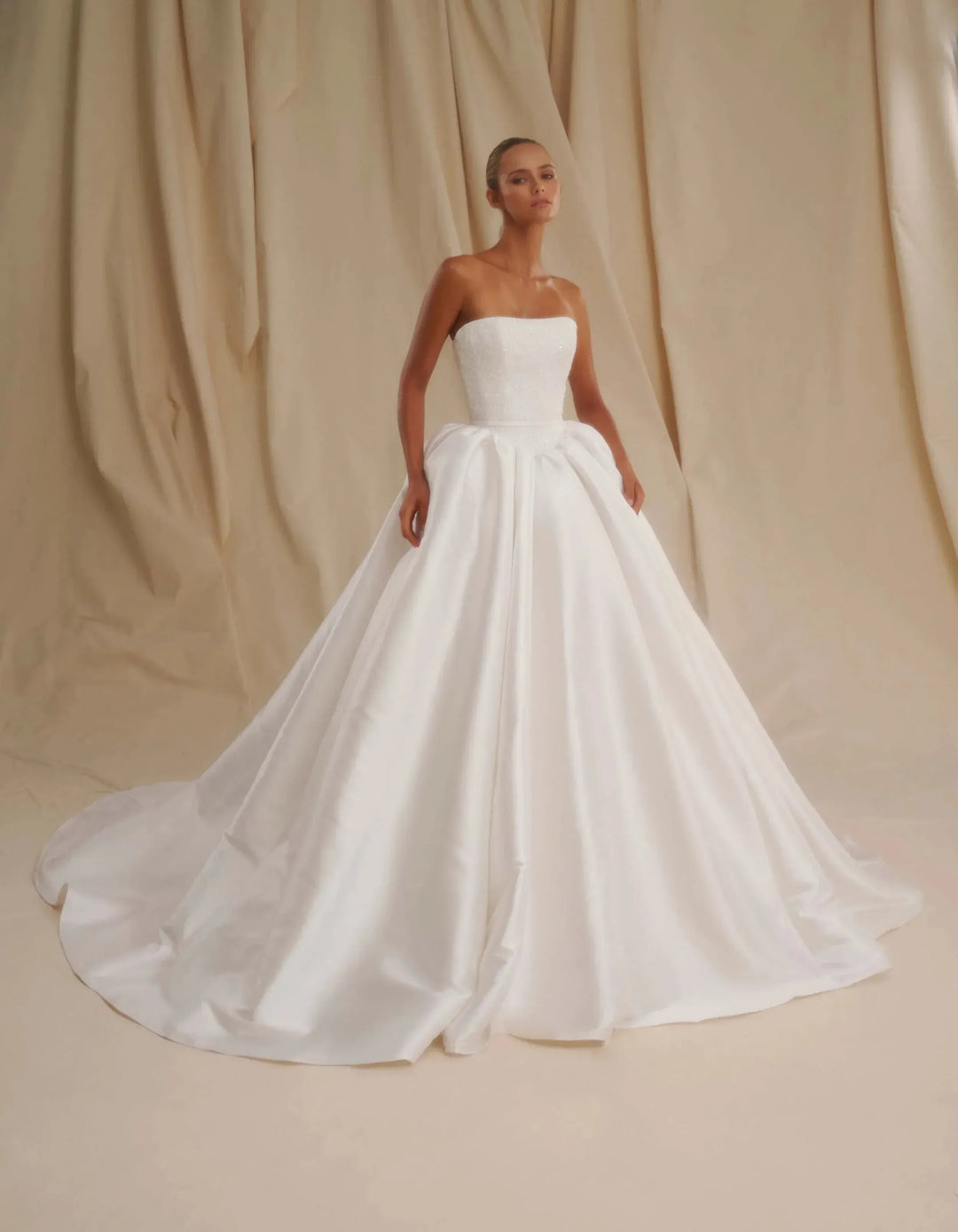 A woman modeling a strapless white ball gown wedding dress with a structured bodice and a full satin train, posed against a draped beige fabric backdrop.