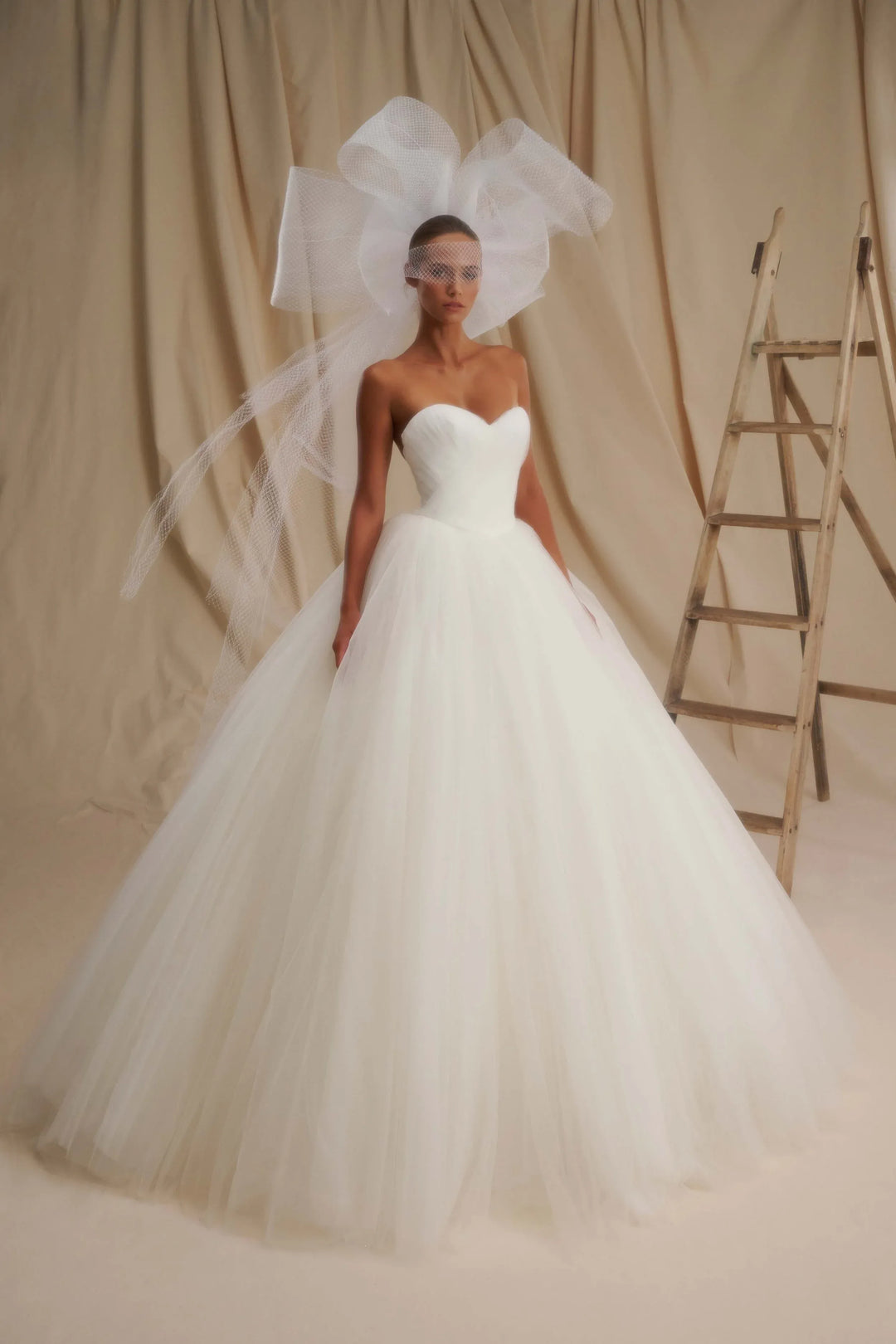 A woman wearing a strapless white wedding gown with a voluminous tulle skirt and a large sheer bow headpiece, standing against a beige draped backdrop with a wooden ladder at the side.