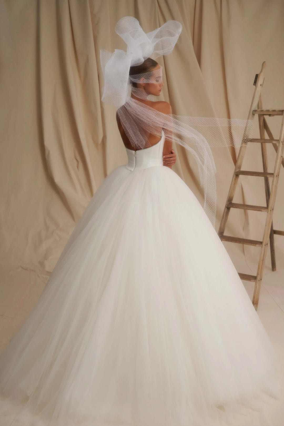 A woman wearing a voluminous white wedding gown with a long sheer veil and a large bow headpiece, photographed against draped beige fabric with a wooden ladder at the side.