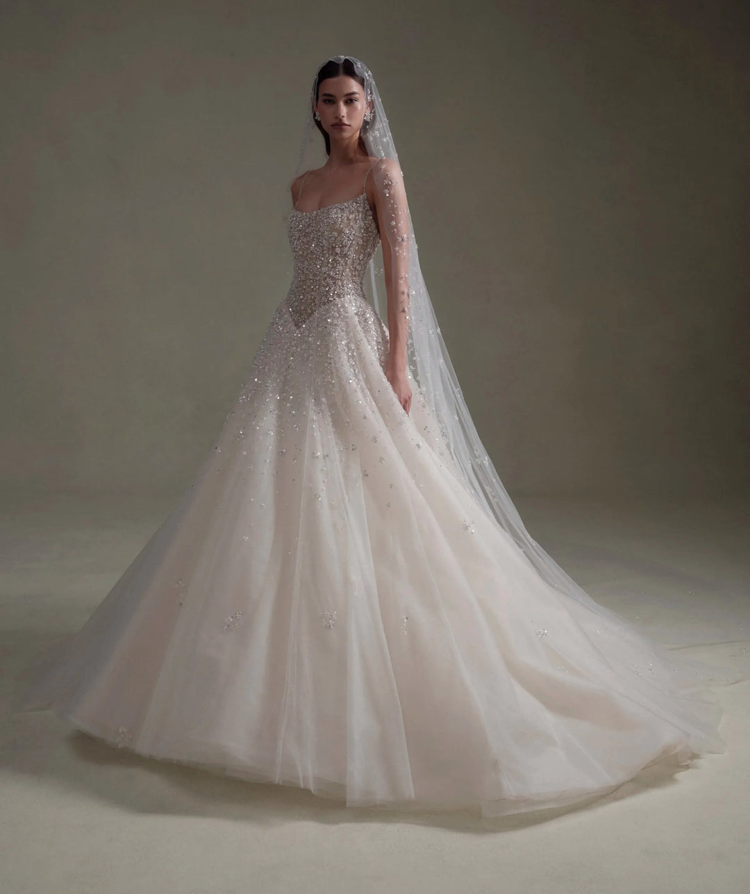 A woman modeling a strapless ball gown wedding dress with a long sheer veil, standing in a studio with a warm beige background.
