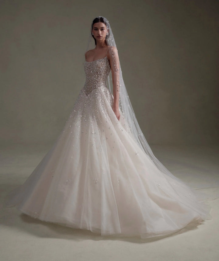 A woman modeling a strapless ball gown wedding dress with a long sheer veil, standing in a studio with a warm beige background.