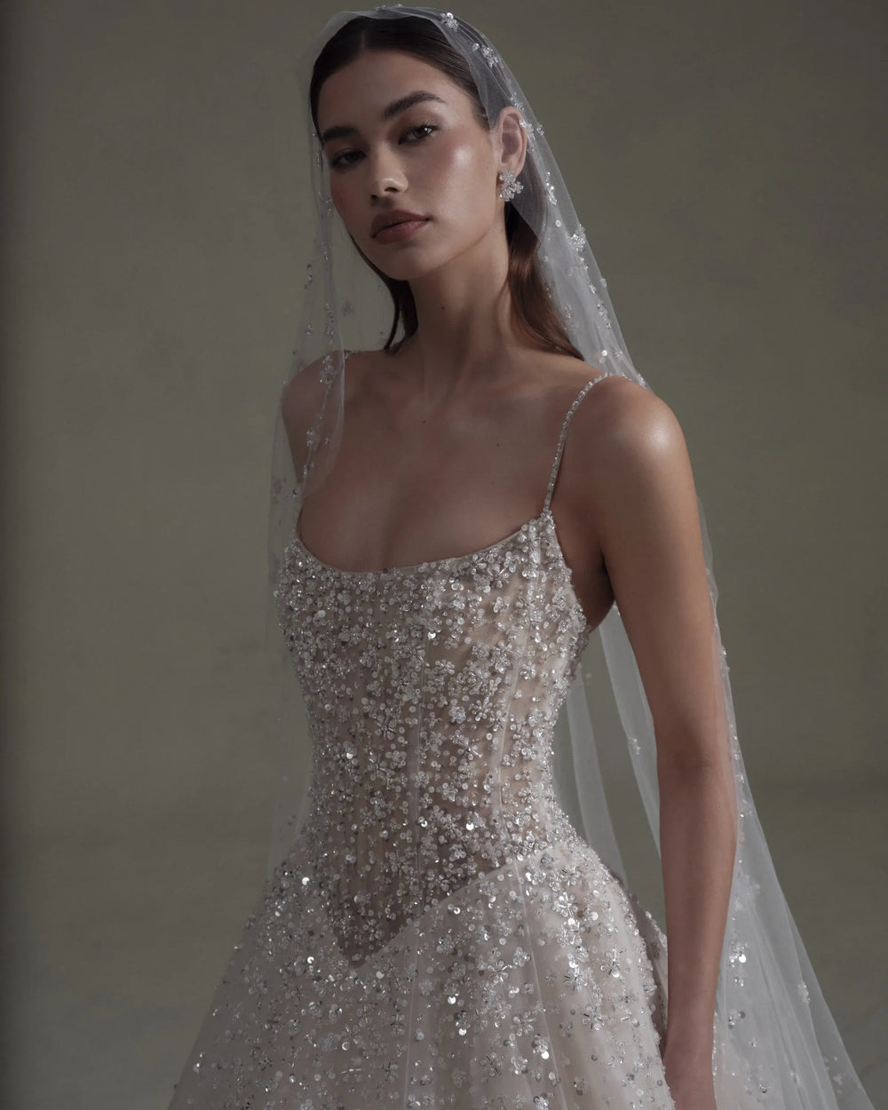 A woman wearing a sparkling silver and white beaded wedding gown with thin straps and a sheer veil, posed against a neutral warm gray background.