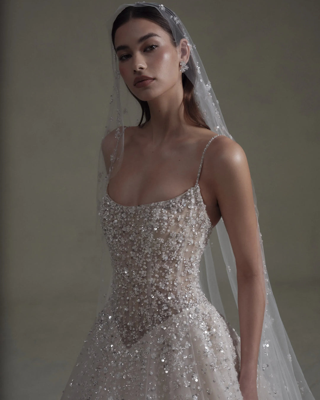 A woman wearing a sparkling silver and white beaded wedding gown with thin straps and a sheer veil, posed against a neutral warm gray background.