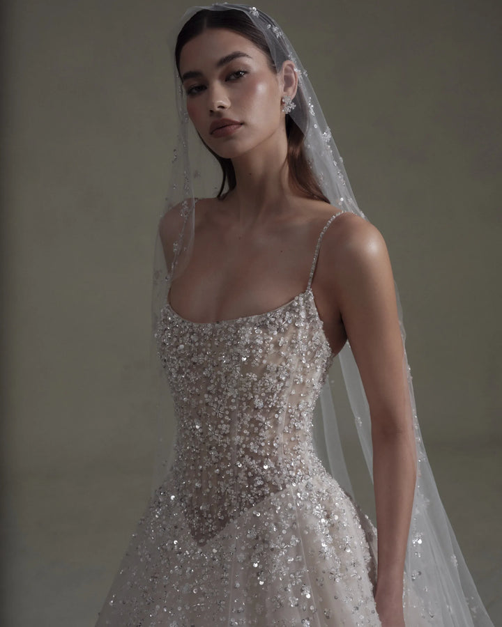 A woman wearing a sparkling silver and white beaded wedding gown with thin straps and a sheer veil, posed against a neutral warm gray background.
