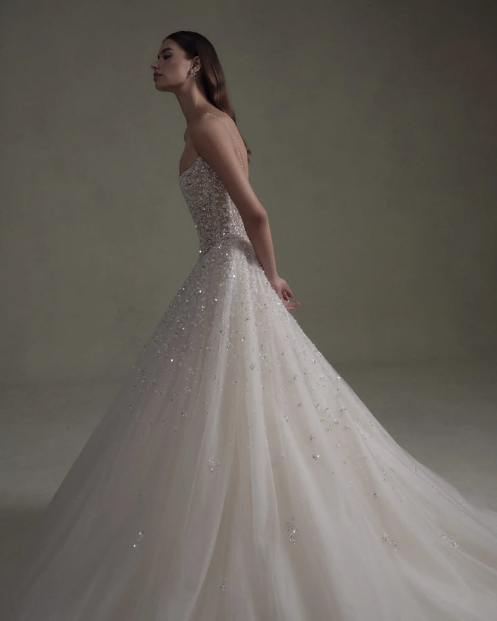 A woman wearing a strapless ball gown wedding dress with intricate beadwork and tulle, photographed in a studio setting against a neutral beige background.